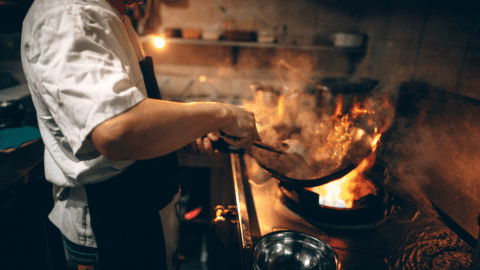 A chef cooking with a pan on a stovetop with flames.