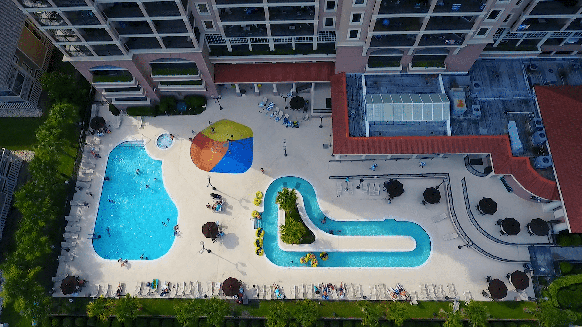 Aerial view of a resort pool area with a swimming pool, water slide, lounge chairs, and umbrellas.