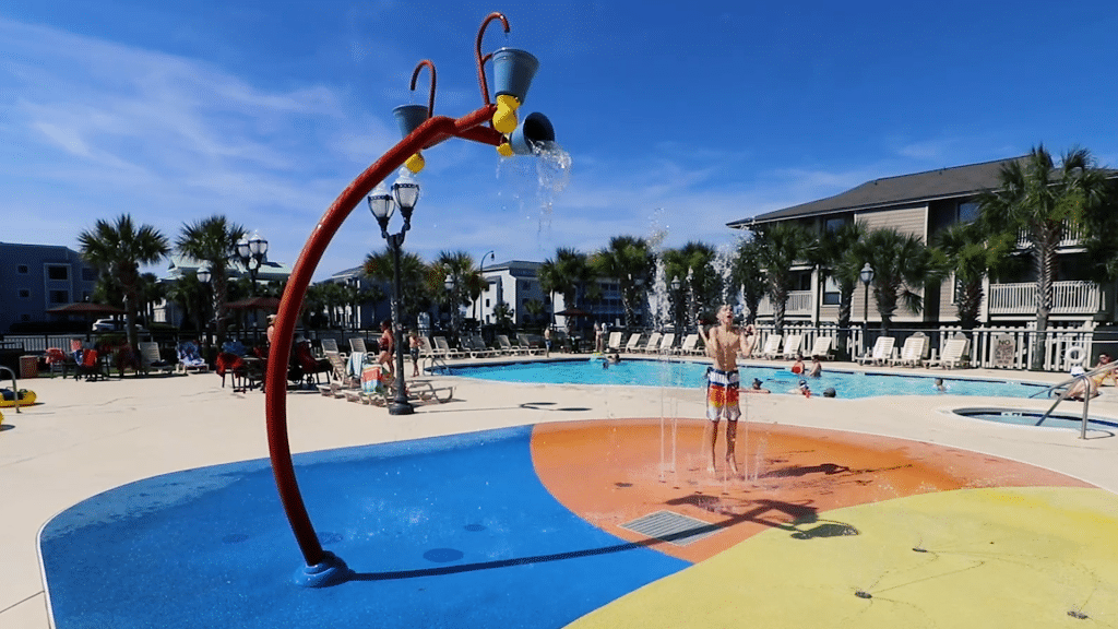 A colorful children's water play area with a large red and blue structure and a pool in the background.